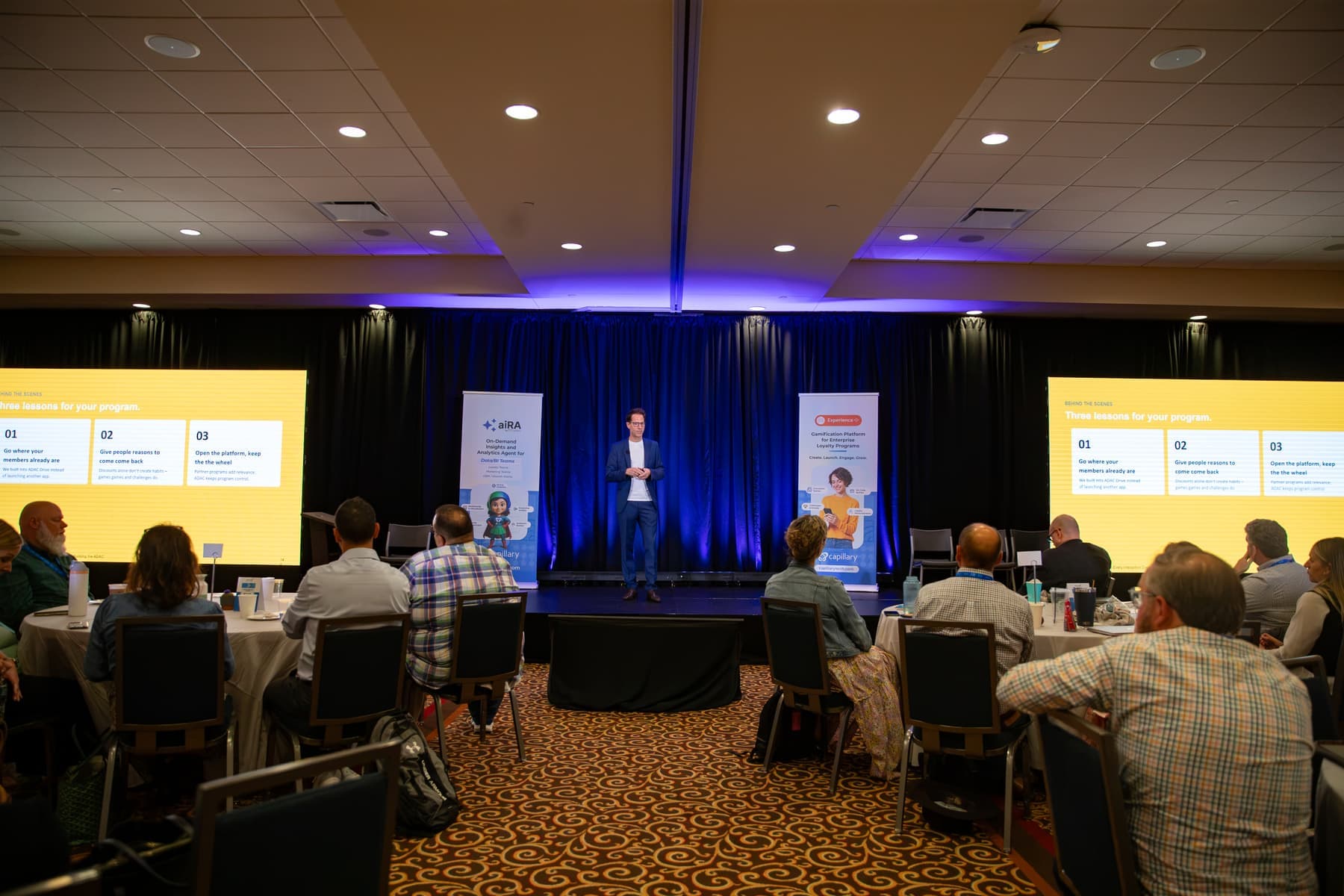 A speaker on stage between two screens showing three lessons for loyalty programs